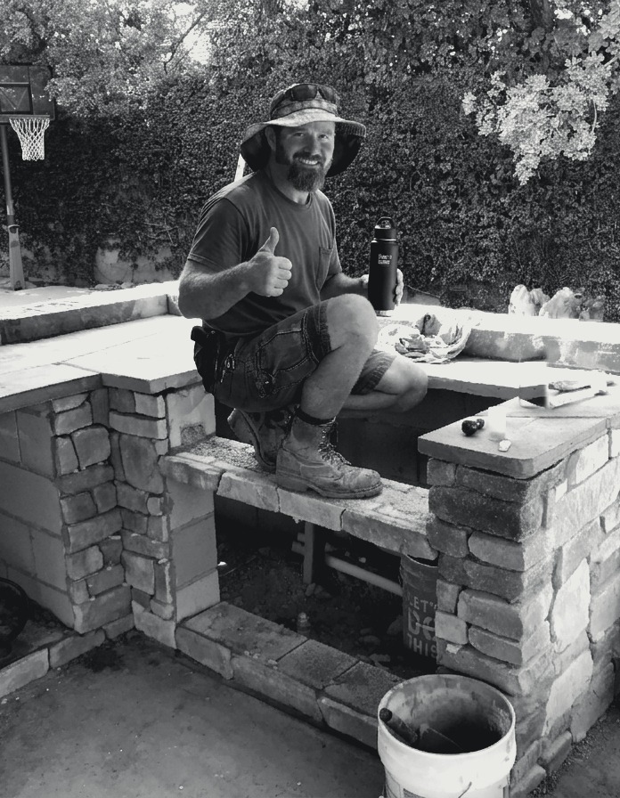James Wright giving a thumbs-up while crouched on a stacked-stone wall he is building
