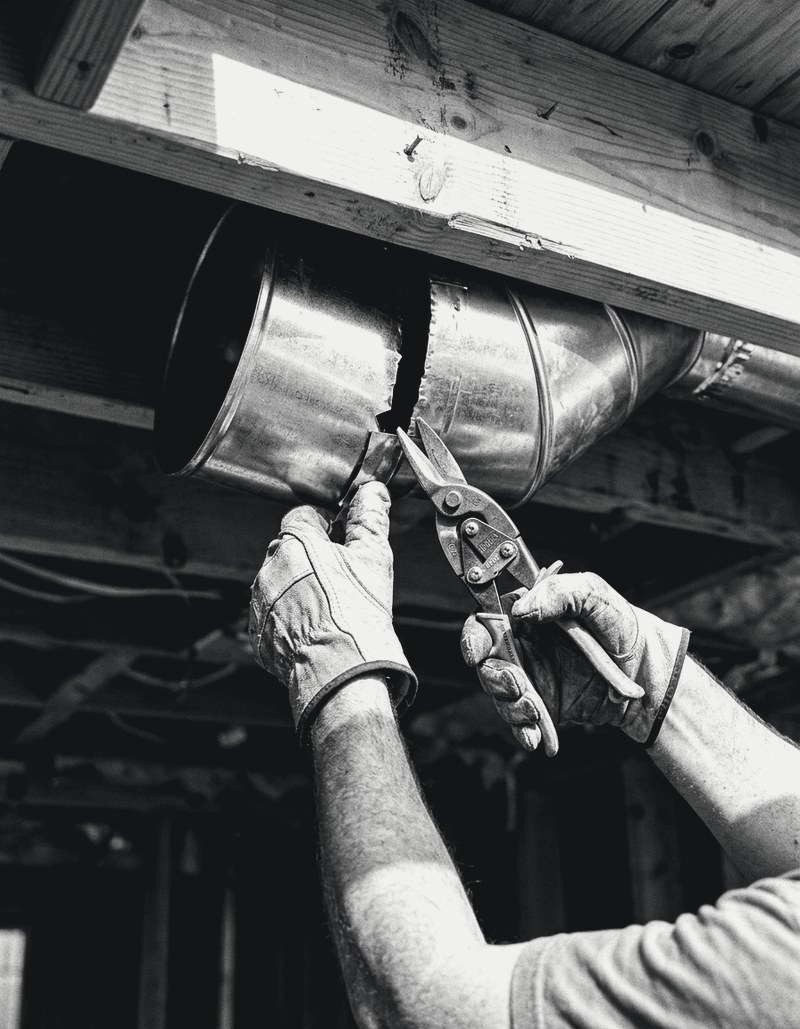 HVAC technician cutting a duct seam with tin snips under floor joists